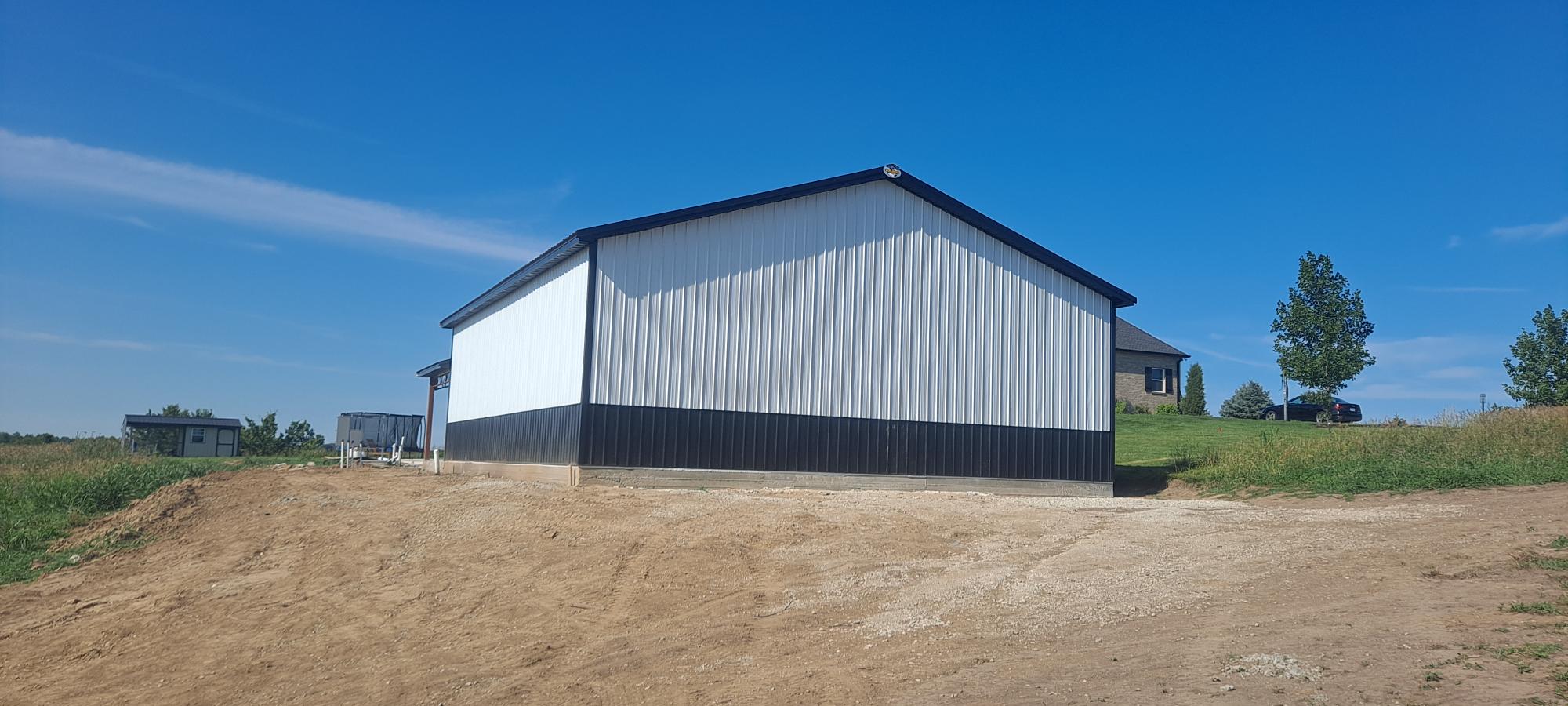 A large white and black metal building sits on a sandy-dirt surface. Green grass and a few trees can be seen on the right side, while the background on the left side features a small structure and a house in the distance under a clear blue sky.