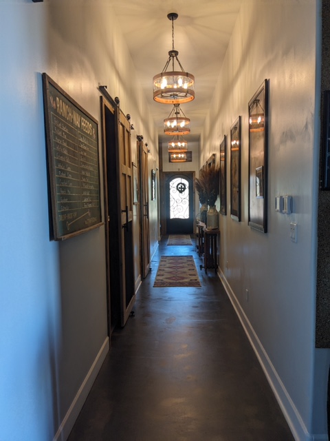 A narrow hallway with wooden floors, decorated with framed pictures on white walls and lit by pendant lights. On the left wall, a large chalkboard with writing is visible. At the end of the hallway, a small table and a door with a decorative glass panel can be seen.