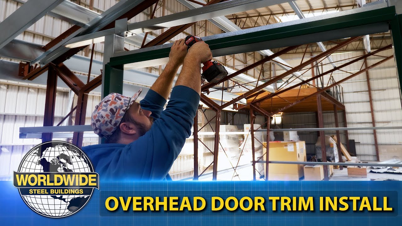 Person in a blue hoodie and hat using a power drill to install overhead door trim in a metal building. Text overlay reads "WORLDWIDE STEEL BUILDINGS" and "OVERHEAD DOOR TRIM INSTALL." Watch our videos for helpful installation tips.